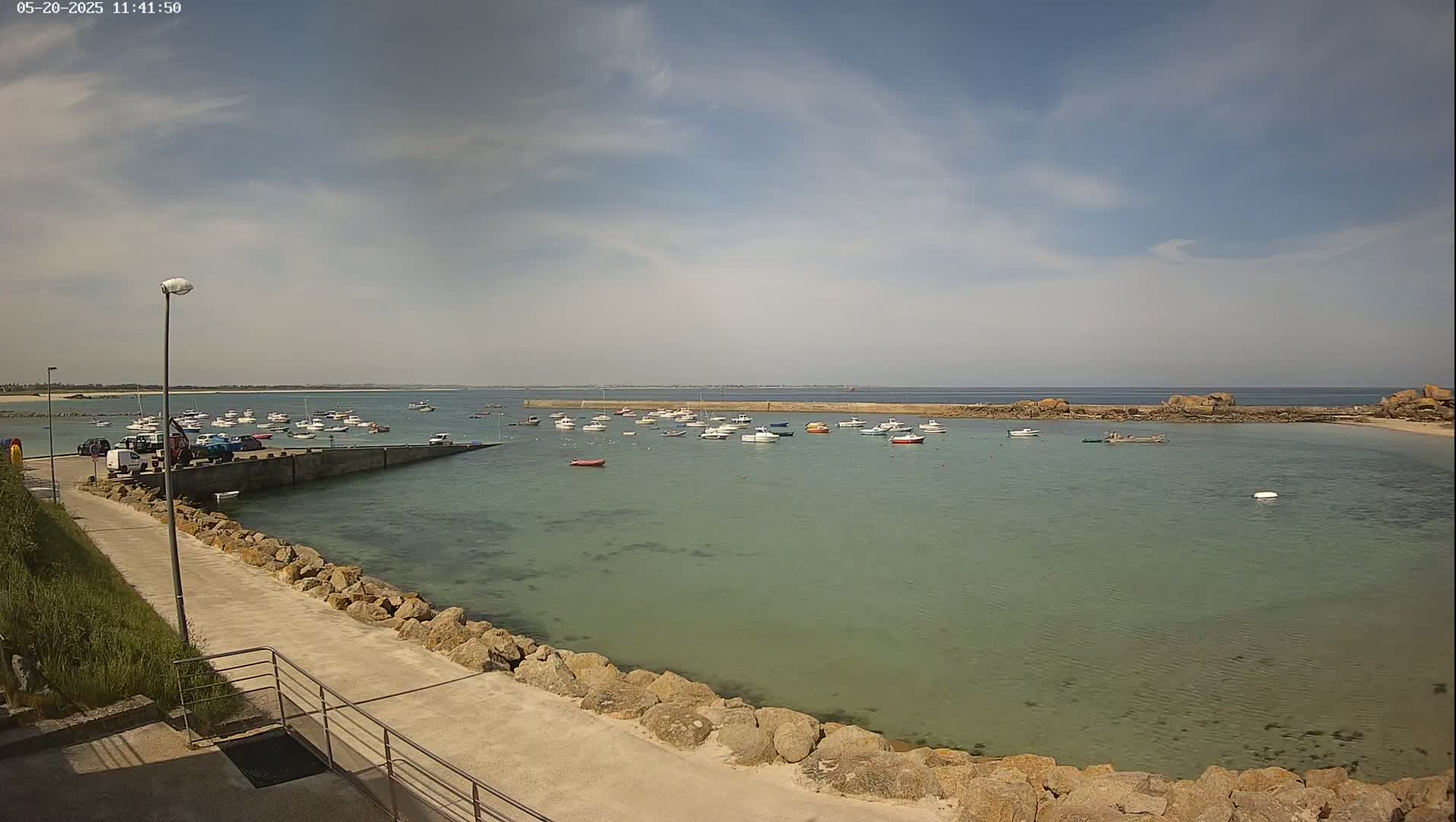 A calm, sunny day reveals a harbor filled with numerous small boats, nestled between a stone walkway and a rocky shoreline extending into a tranquil sea.