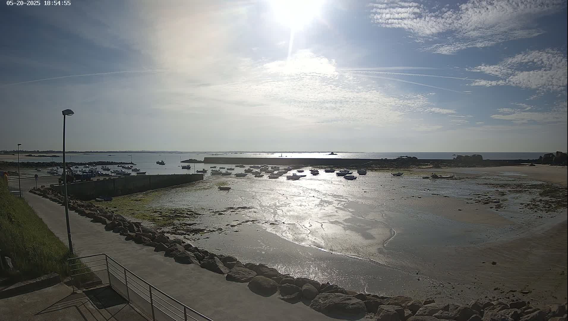 A mostly sunny day reveals a harbor filled with numerous small boats at low tide, surrounded by rocky shores and calm water reflecting sunlight.