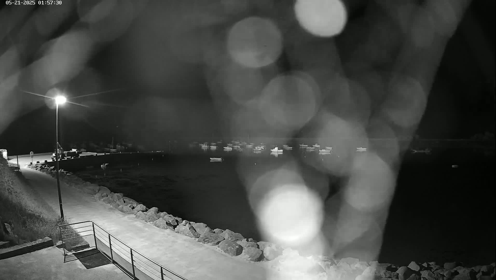 A nighttime, rain-blurred view of a harbor with numerous small boats at anchor, a paved walkway leading to a lamplit area, and a rocky shoreline.