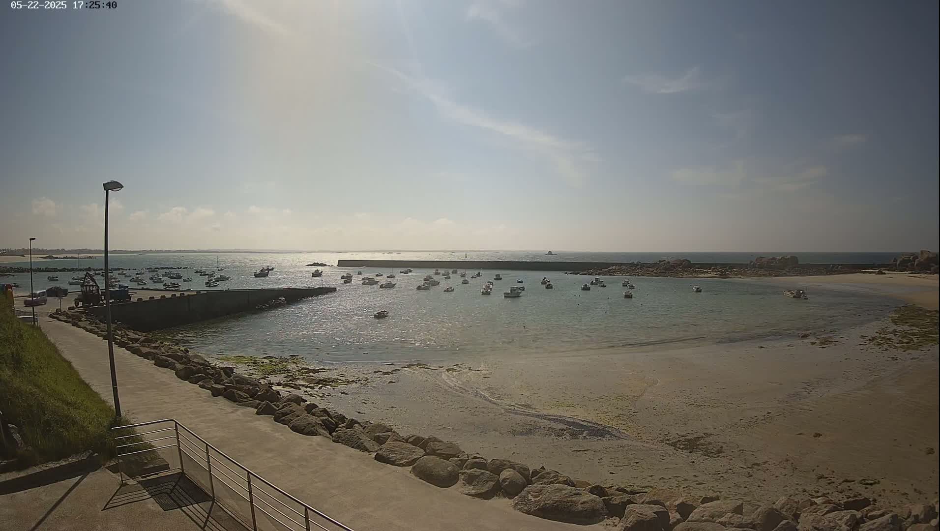 A sunny day at a harbor shows numerous small boats in calm water, a sandy beach, and a paved walkway leading to the water.
