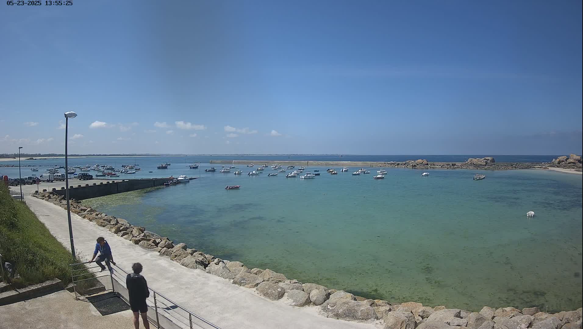 A calm, clear day shows a harbor filled with numerous small boats, a paved walkway with two people, and rocky shores under a bright blue sky.
