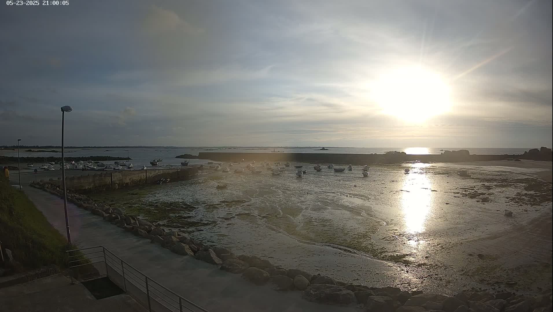 A harbor filled with small boats at low tide is bathed in the golden light of a setting sun under a partly cloudy sky.