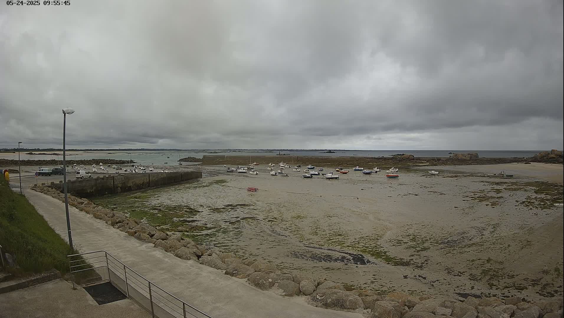 A mostly cloudy day reveals a small harbor filled with numerous small boats at low tide, surrounded by rocks and a paved walkway.