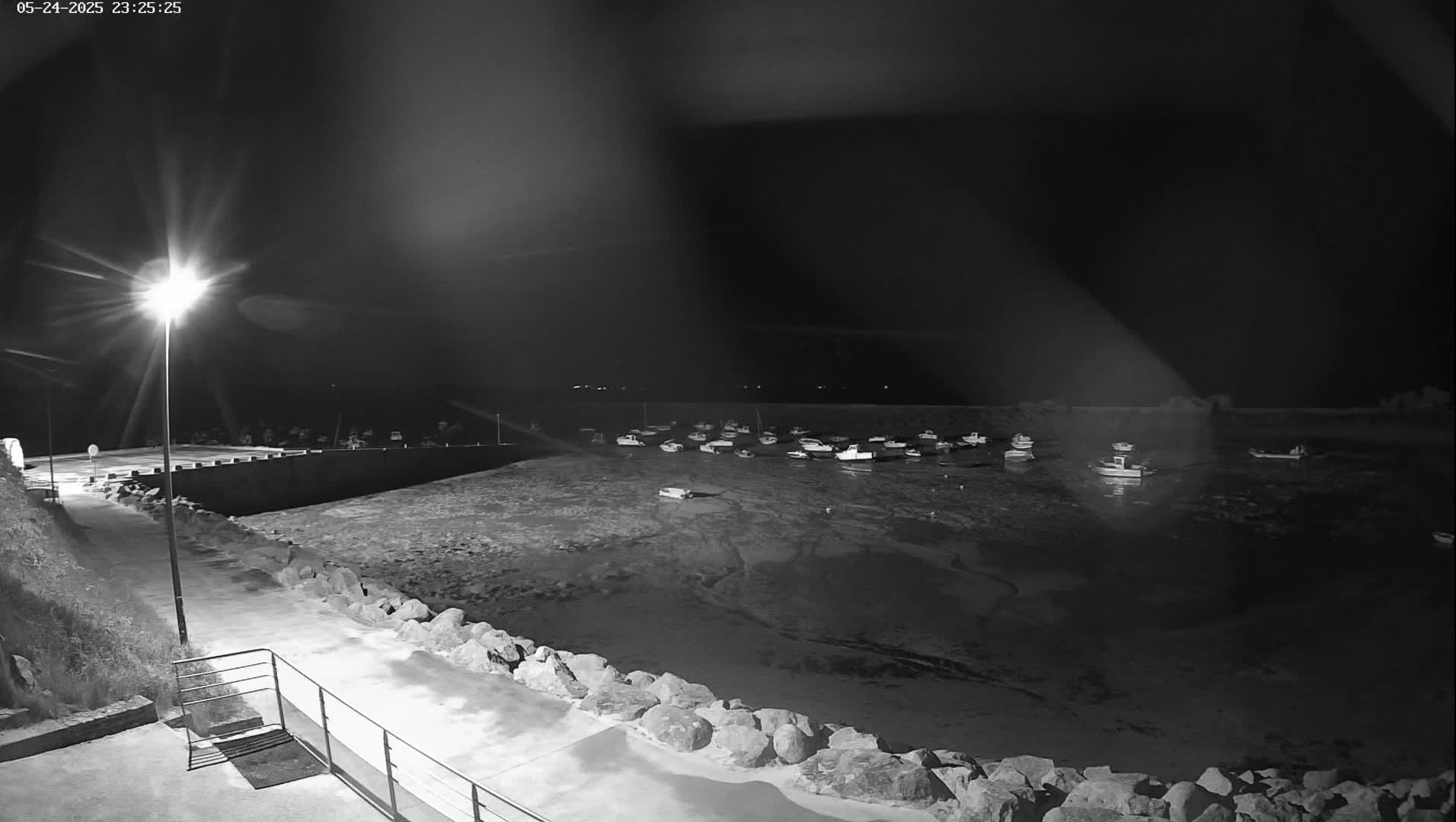 A nighttime, low-light view shows a harbor with numerous small boats at low tide, illuminated by a single bright streetlight on a stone walkway.
