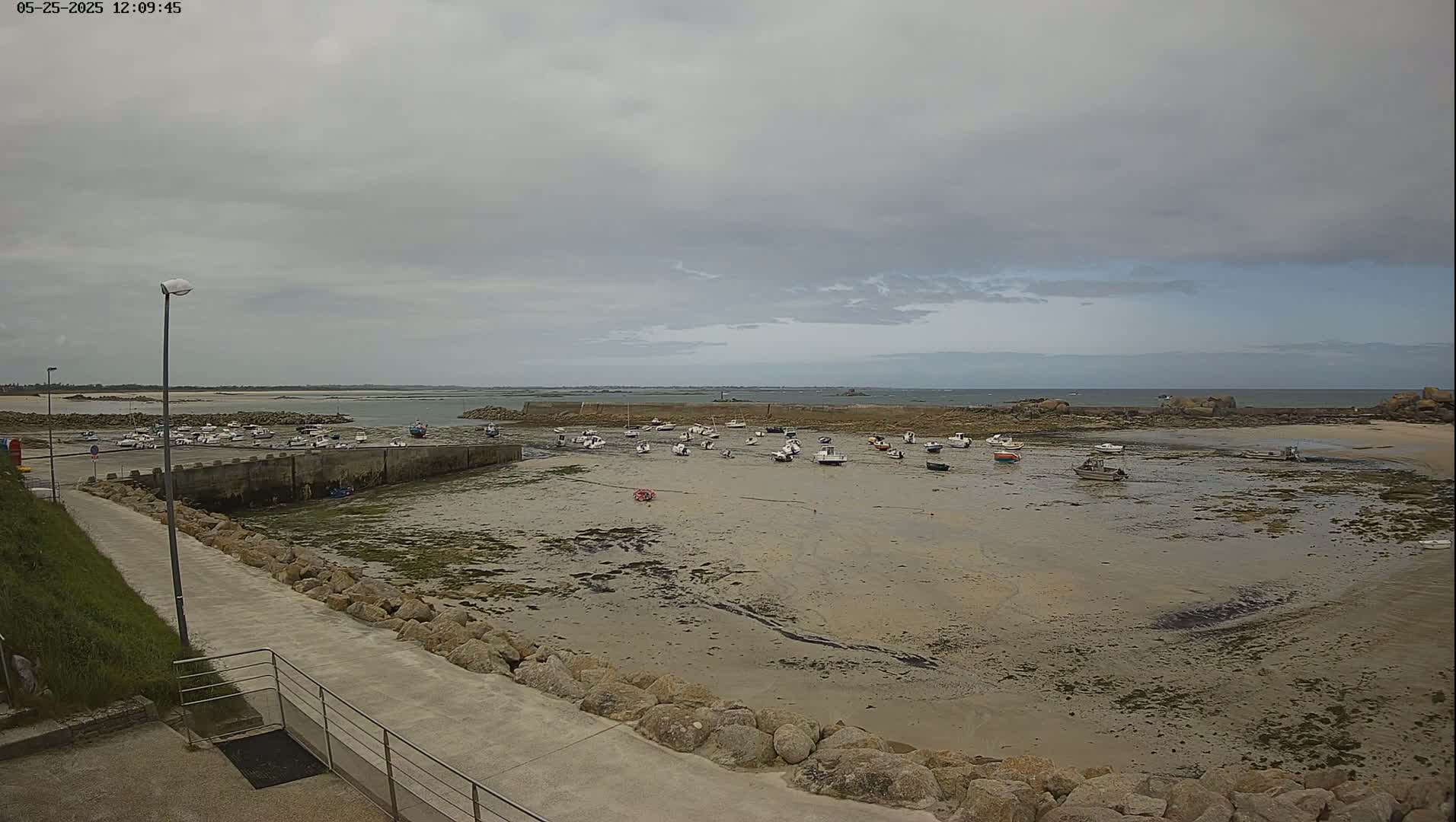 A mostly cloudy day reveals a small harbor with numerous boats at low tide, surrounded by rocky shores and calm water extending to the horizon.