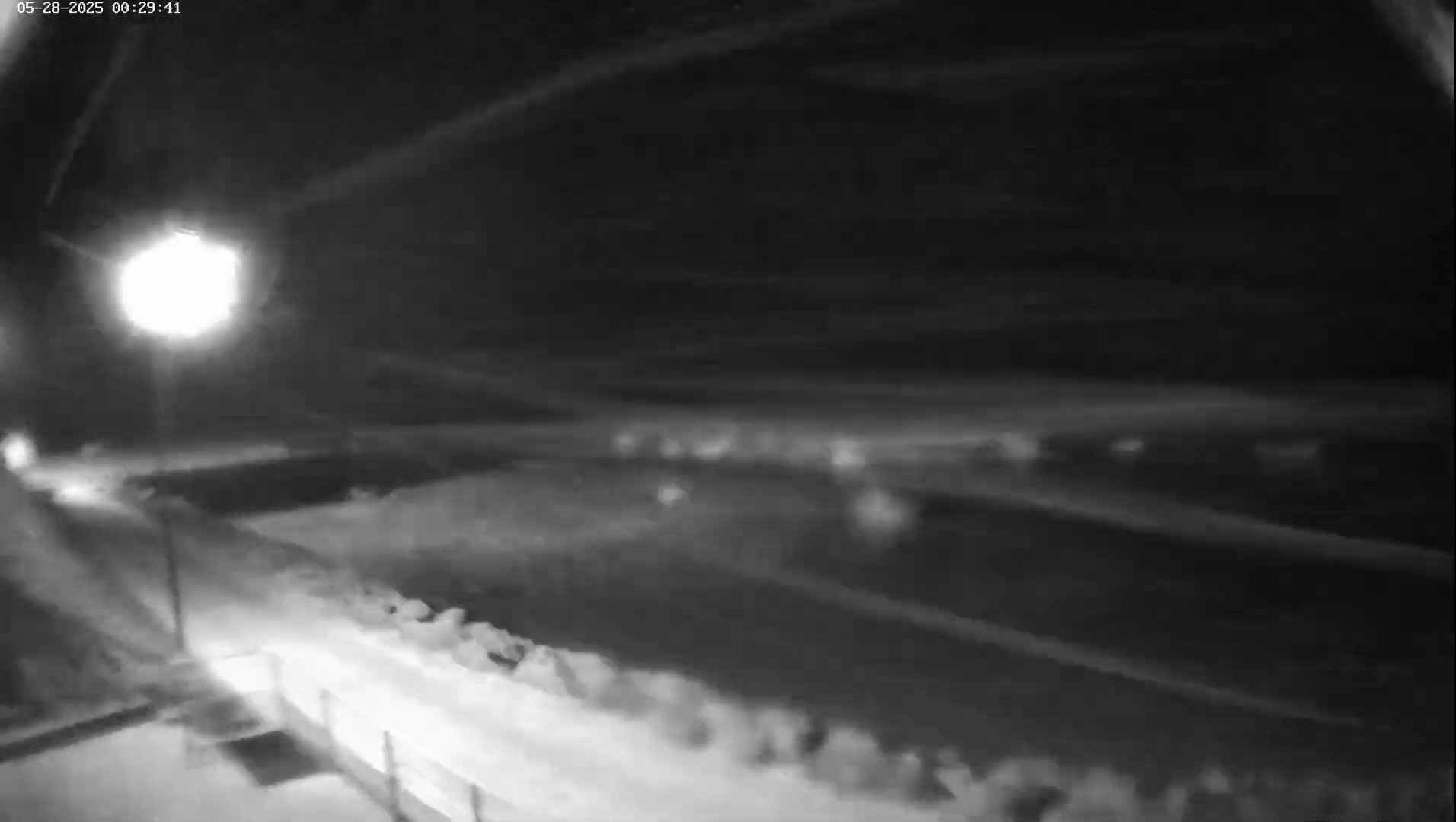 A nighttime, snowy scene shows a road under a cloudy sky illuminated by a bright streetlight.