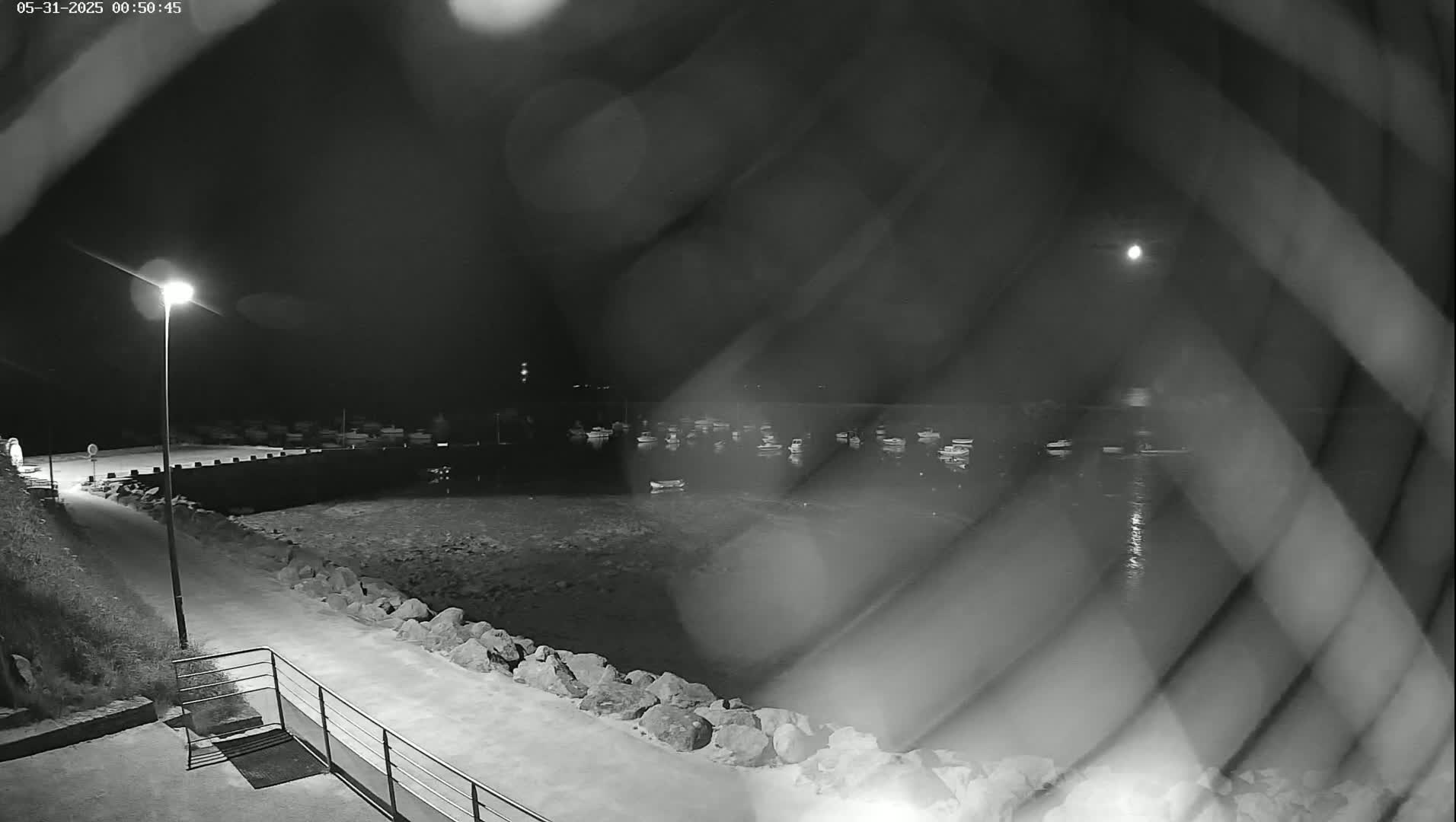 A nighttime, low-light view through a fence shows a harbor with numerous boats at rest, a paved walkway, and a single lit lamppost.
