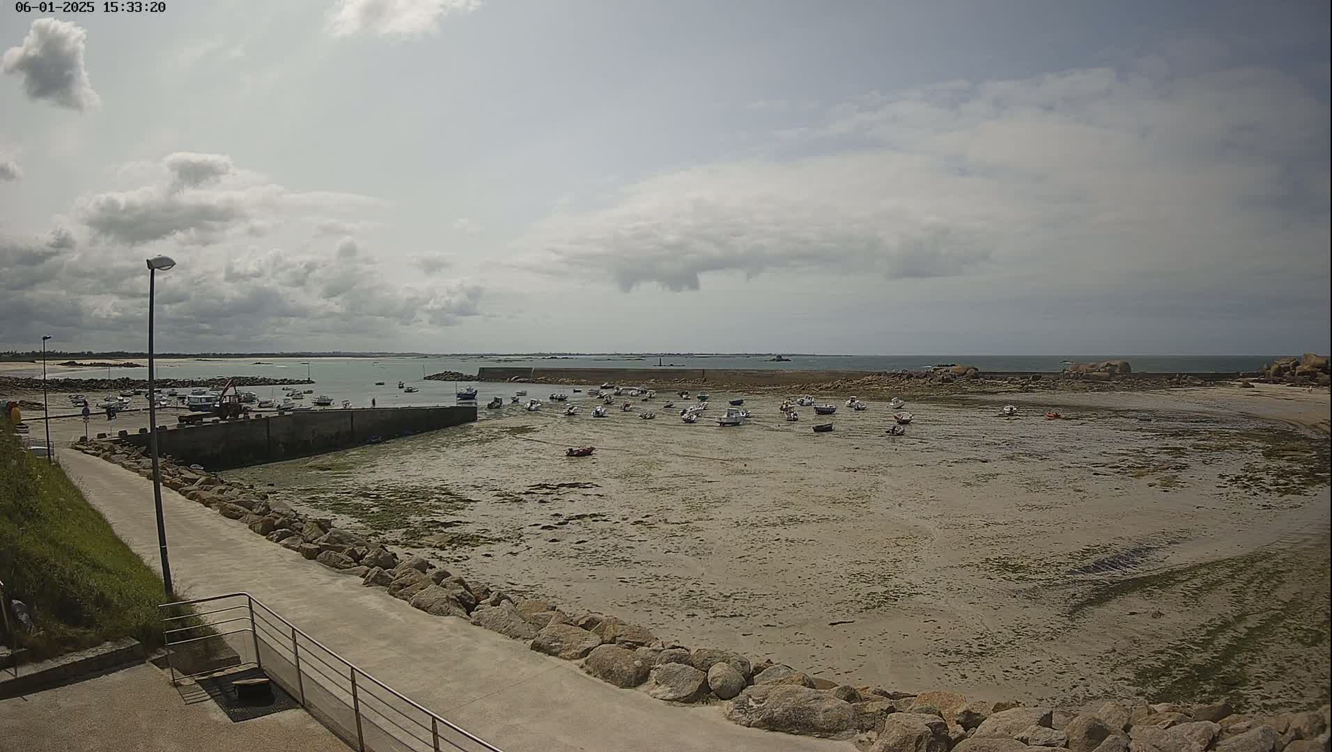A mostly sunny day reveals a harbor filled with numerous small boats resting on exposed mudflats, surrounded by a rocky shoreline and a calm body of water extending to the horizon.