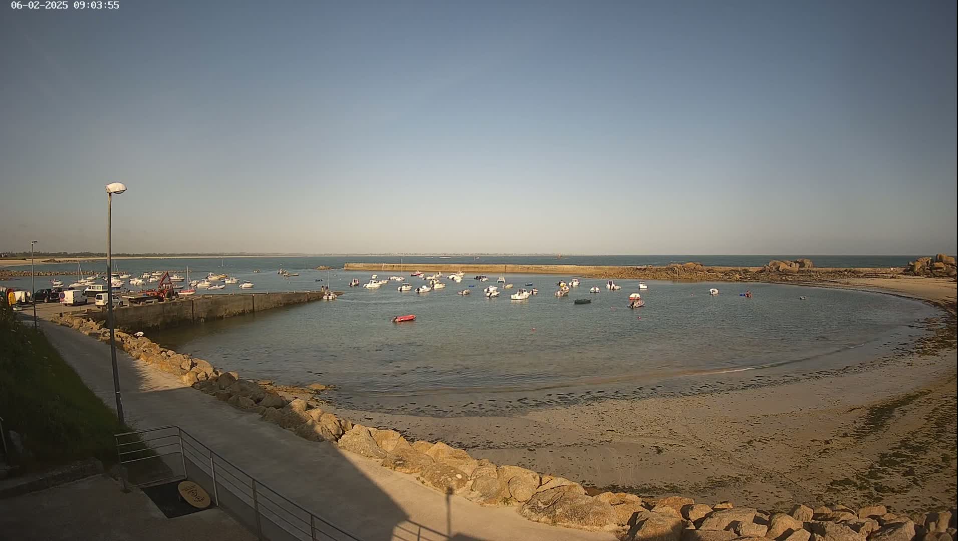 A calm, sunny day reveals a small harbor filled with numerous boats, nestled within a rocky coastline and sandy beach.
