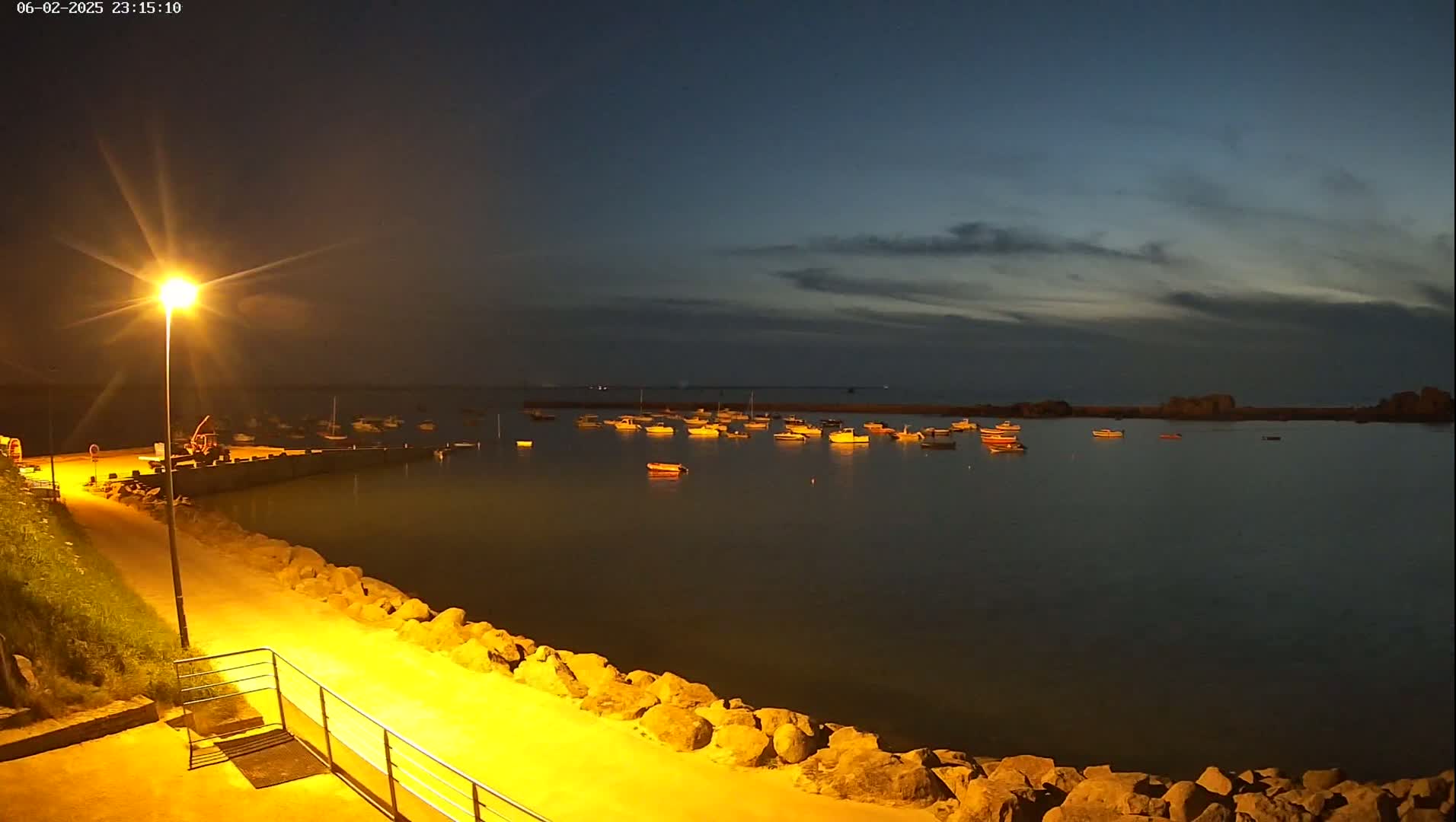 A nighttime view of a calm harbor with numerous small boats moored, illuminated by a single streetlamp on a rocky shoreline under a partly cloudy sky.