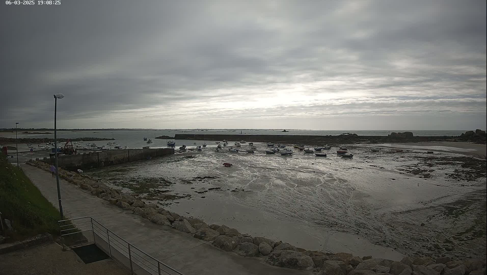 A harbor filled with numerous small boats sits on a calm, overcast day at low tide, with a rocky shoreline and a calm sea extending to the horizon.