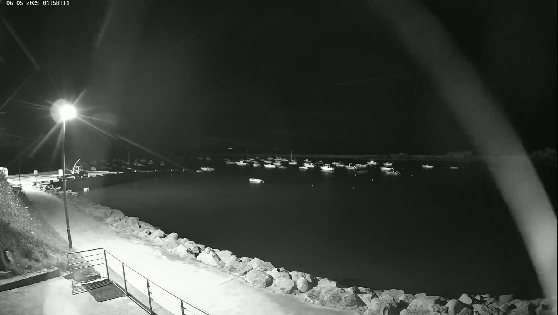 A nighttime view of a harbor filled with numerous boats, illuminated by a single lamppost on a rocky shoreline.