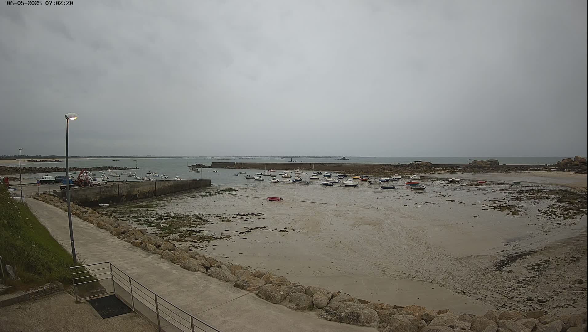 A mostly overcast day reveals a harbor filled with numerous small boats at low tide, with a walkway and a lamppost visible in the foreground.