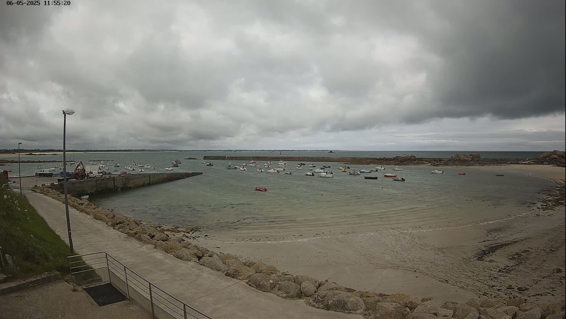 A small harbor filled with numerous boats is seen under an overcast sky, with a sandy beach visible in the foreground.