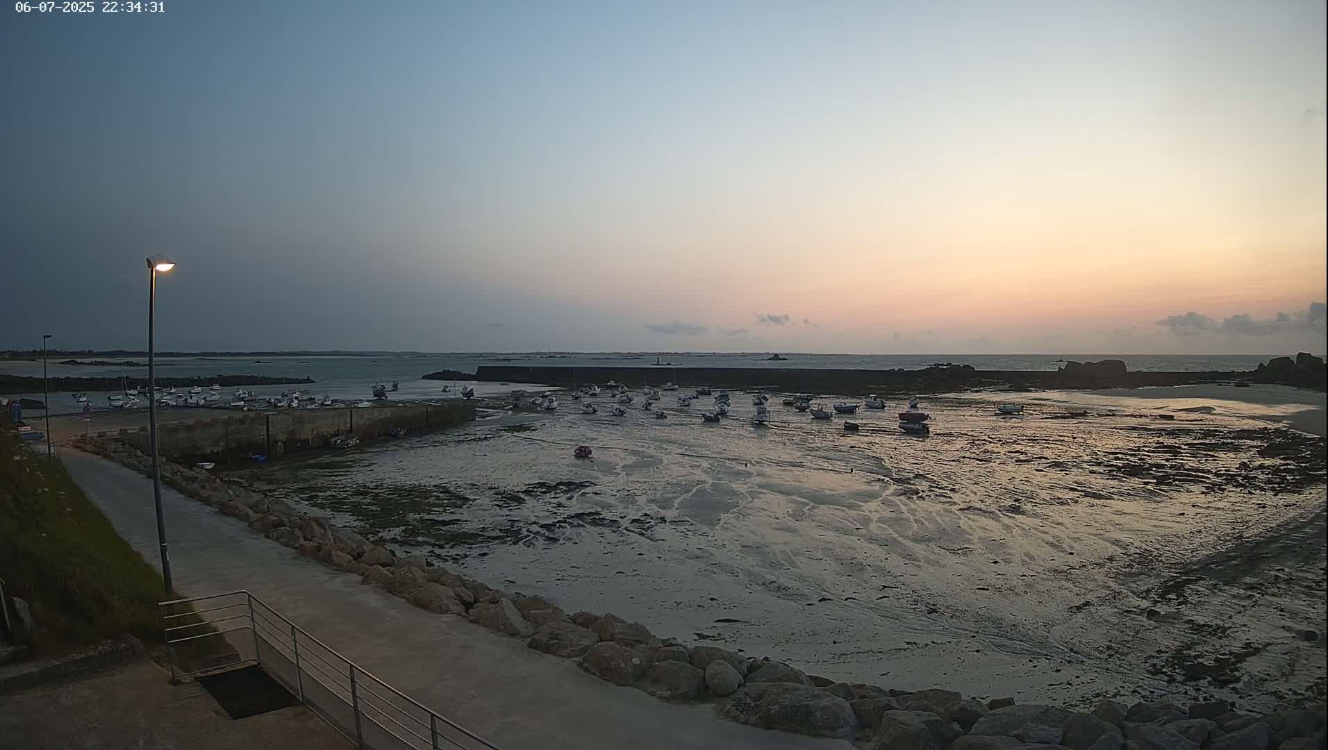 Numerous small boats are moored in a harbor at low tide during a calm sunset.