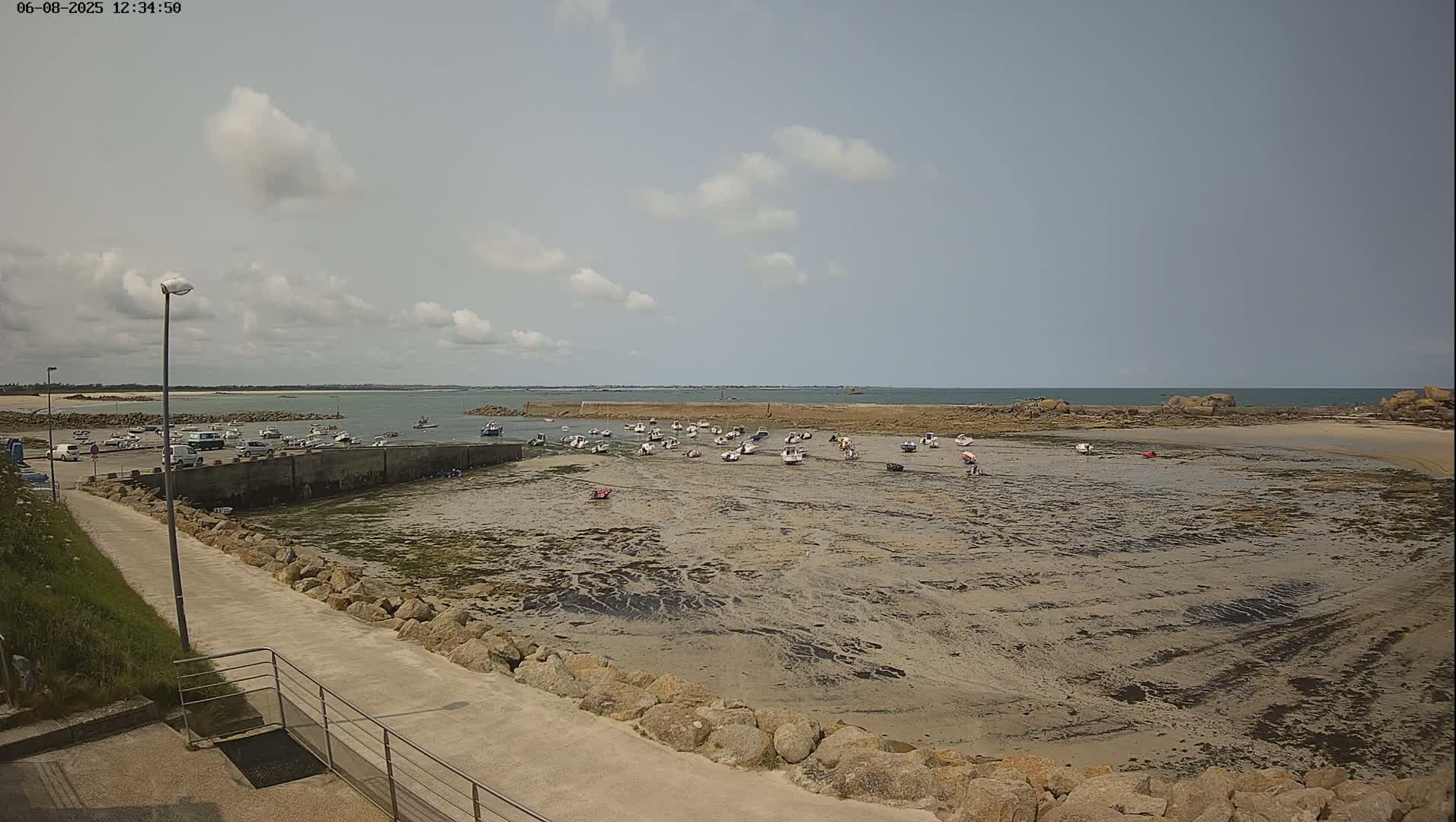 A mostly sunny day shows a harbor filled with numerous small boats resting on a tidal flat, with a calm sea extending to a distant rocky shoreline.