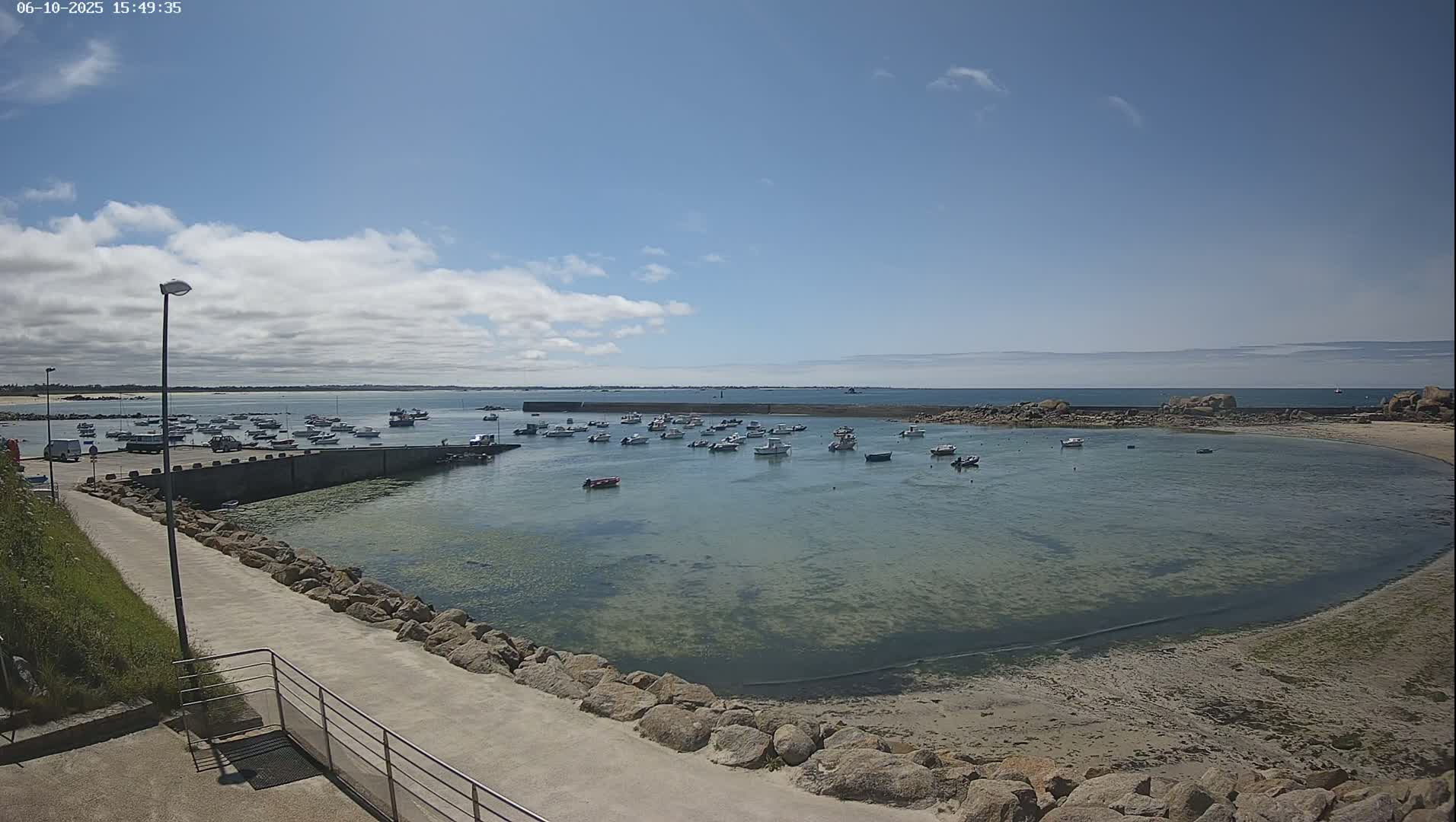 A calm, sunny day shows numerous small boats in a sheltered harbor, surrounded by rocky shores and a clear, shallow sea.