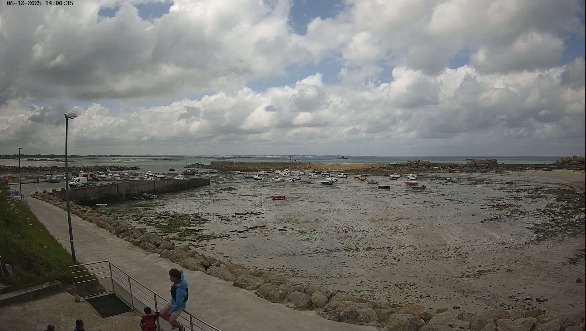 A partly cloudy day reveals a harbor scene with numerous small boats resting on a mudflat at low tide, a few people standing near a walkway, and a calm sea in the distance.