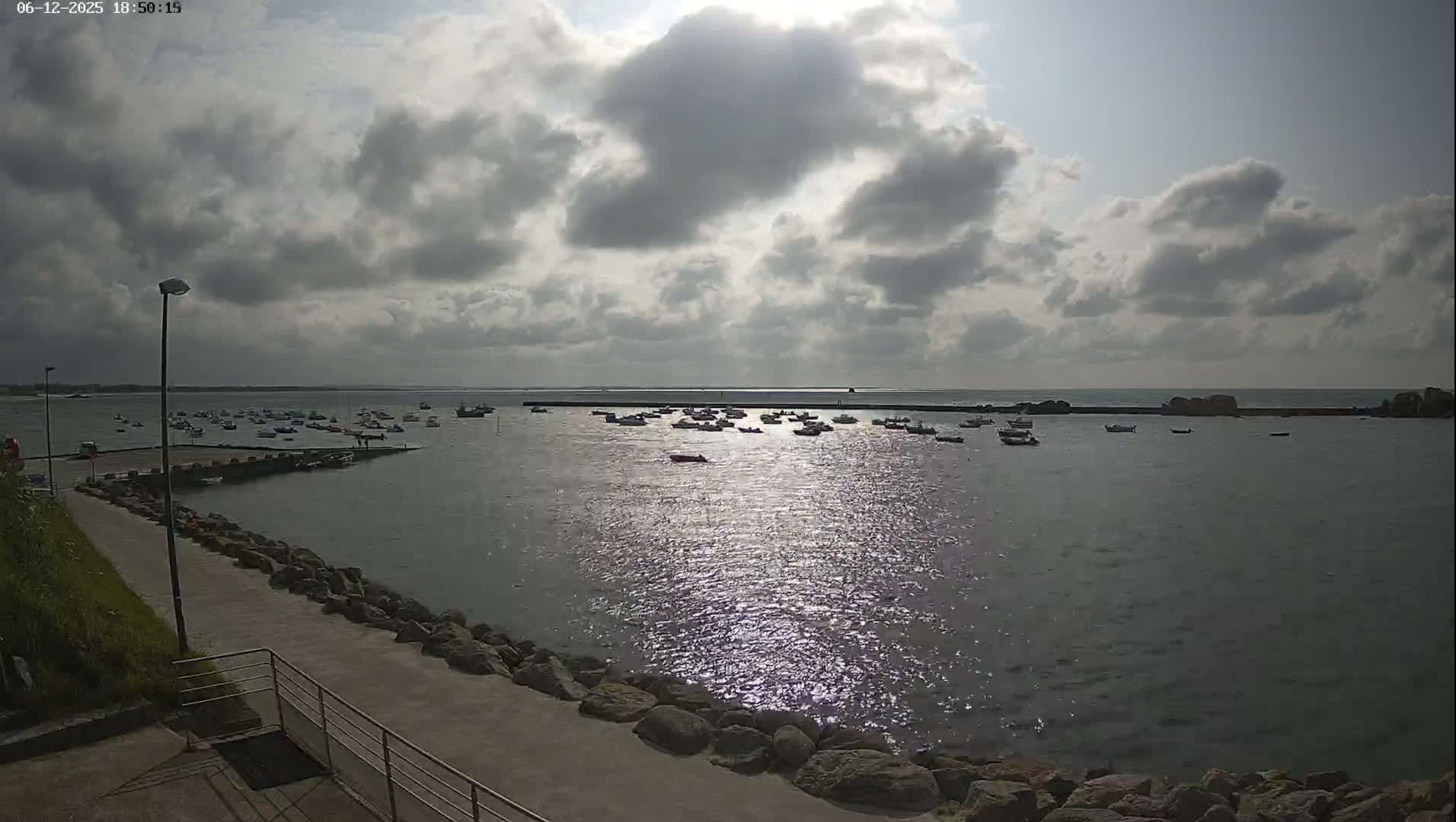 A partly cloudy day reveals a harbor filled with numerous small boats, a rocky shoreline, and a paved walkway leading to the water.