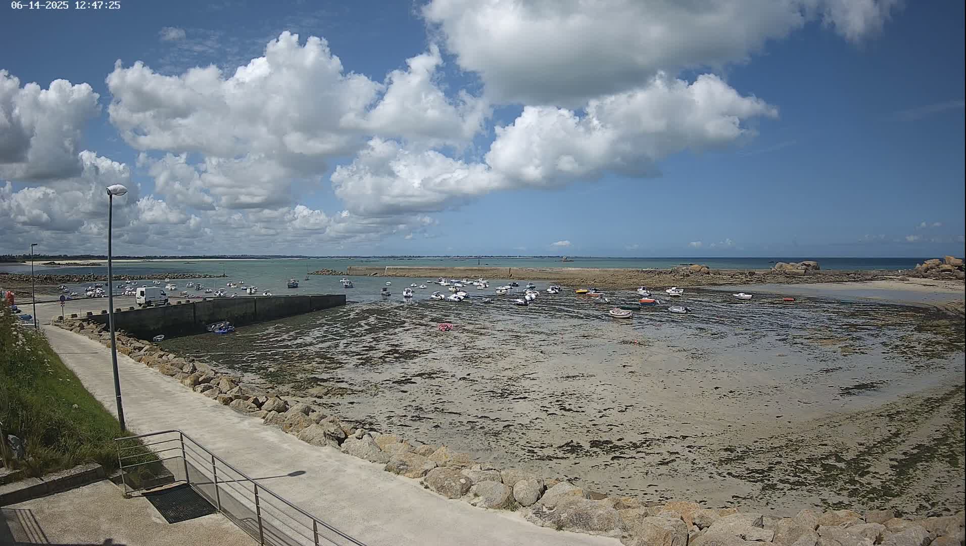 A mostly sunny day reveals a harbor filled with numerous small boats at low tide, surrounded by rocky shorelines and a calm ocean under a partly cloudy sky.