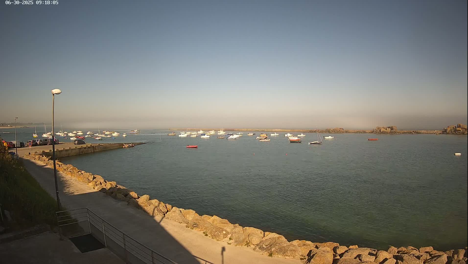 A calm harbor filled with numerous small boats is seen on a hazy, sunny day from a rocky vantage point.