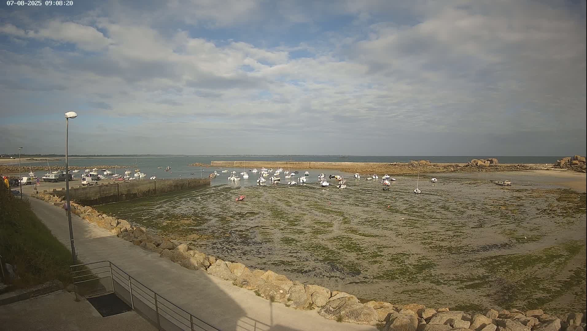 A small harbor filled with numerous boats sits partially exposed at low tide under a partly cloudy sky.