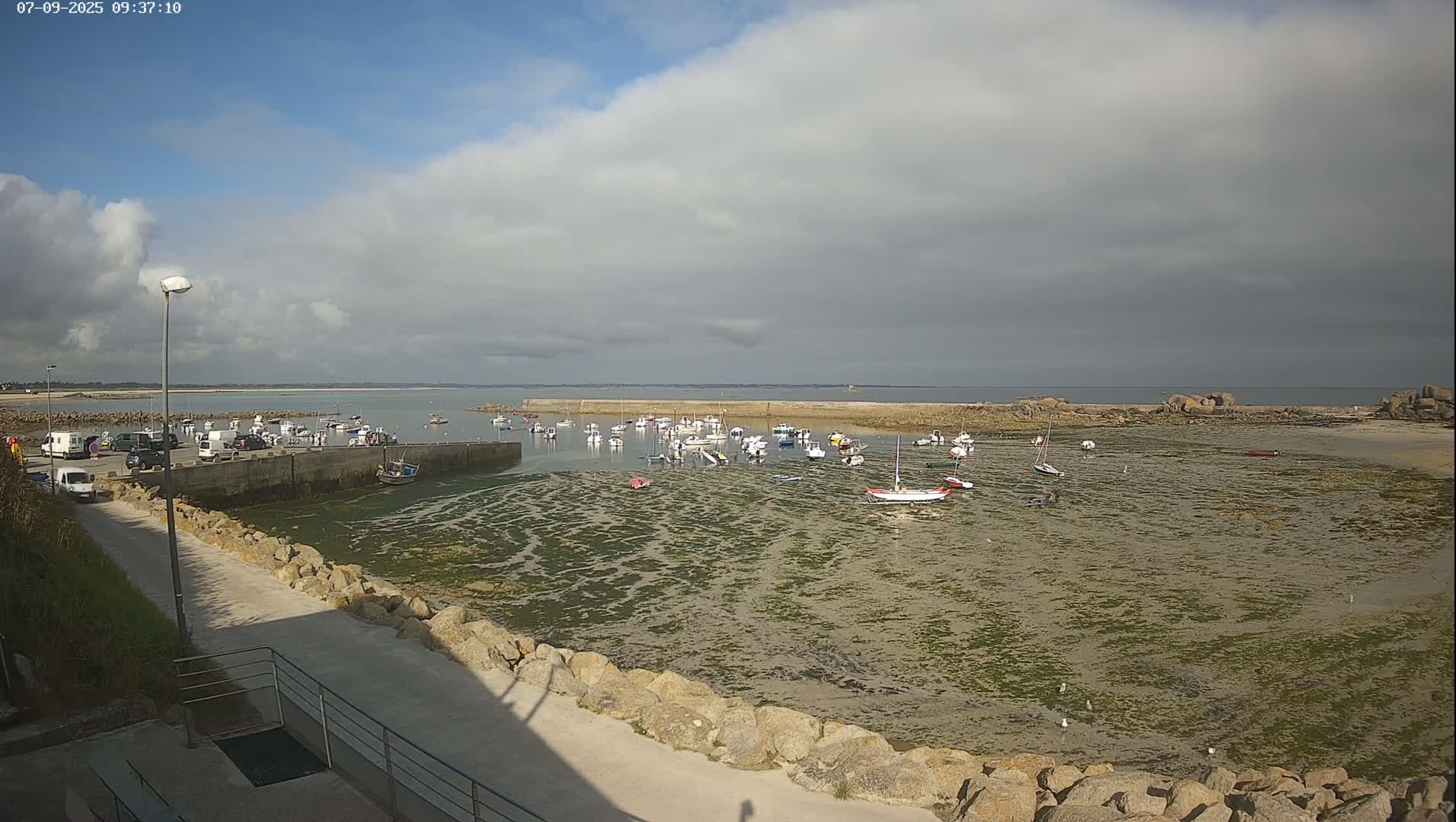 A mostly cloudy day reveals a harbor filled with numerous small boats at low tide, partially exposed seafloor covered with seaweed, and a parking lot with vehicles nearby.