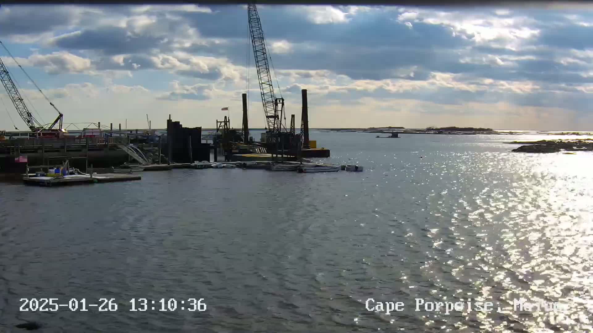 A harbor scene under a partly cloudy sky shows construction equipment and boats near a pier with calm water glistening in the sunlight.