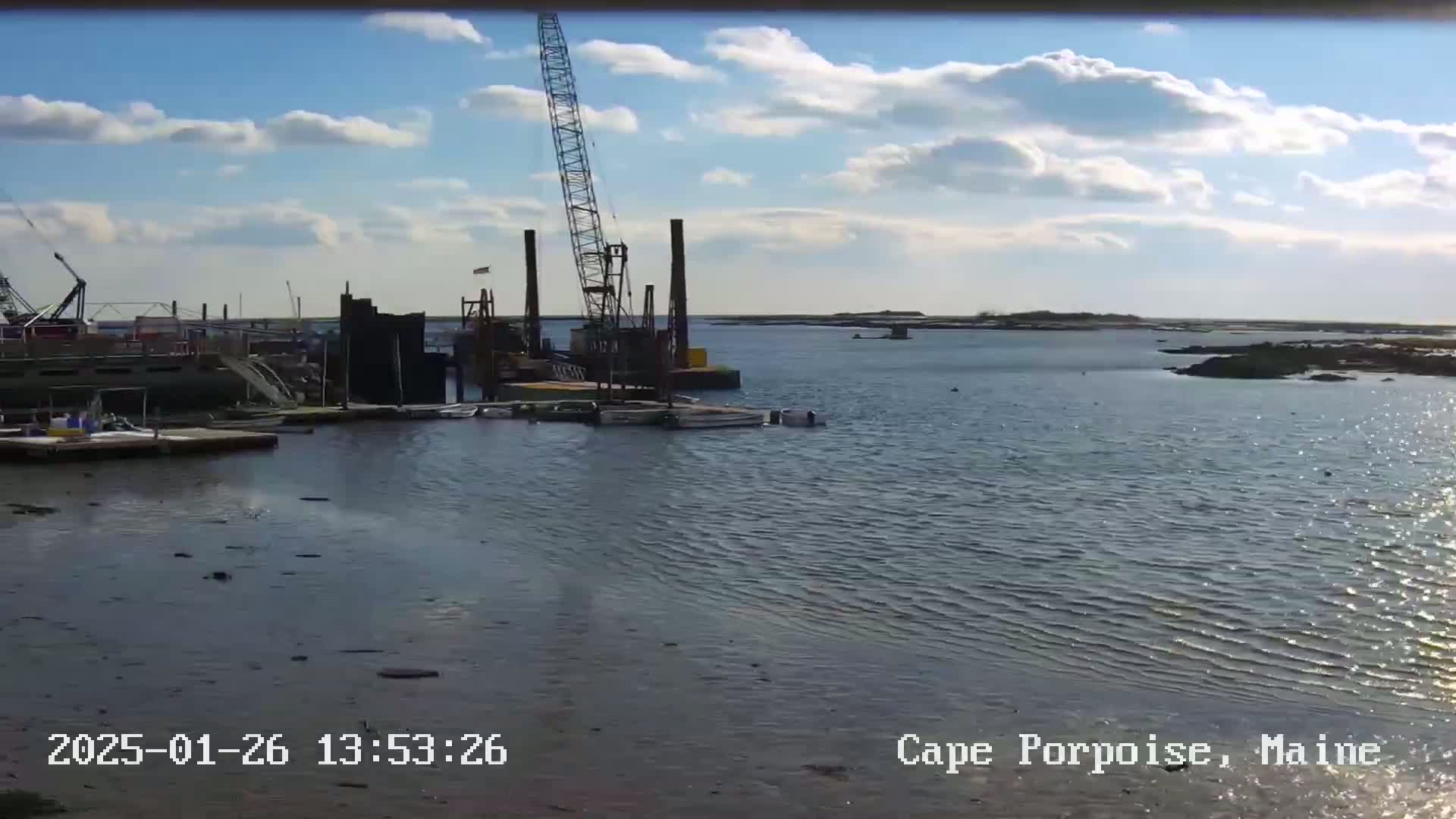 A partly cloudy day shows a calm body of water with a construction crane and several boats near a dock.