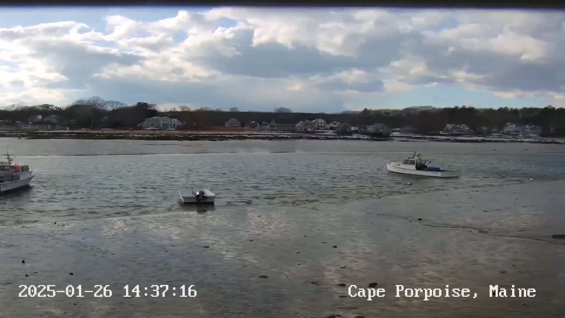 Three small boats are in calm, shallow water near a shoreline with houses under a partly cloudy sky.