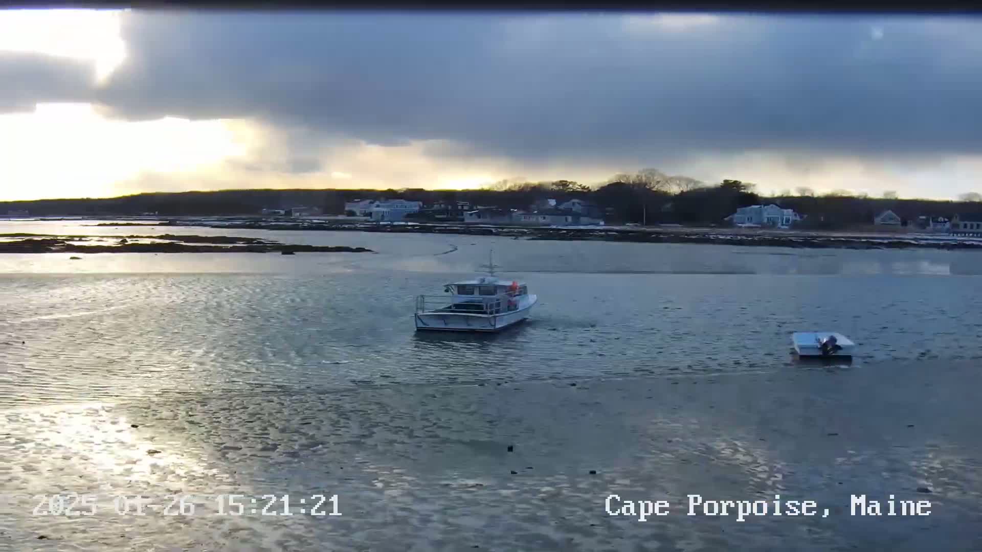 A passenger boat and a smaller boat are on calm water in front of a shoreline with houses under a partly cloudy sky.