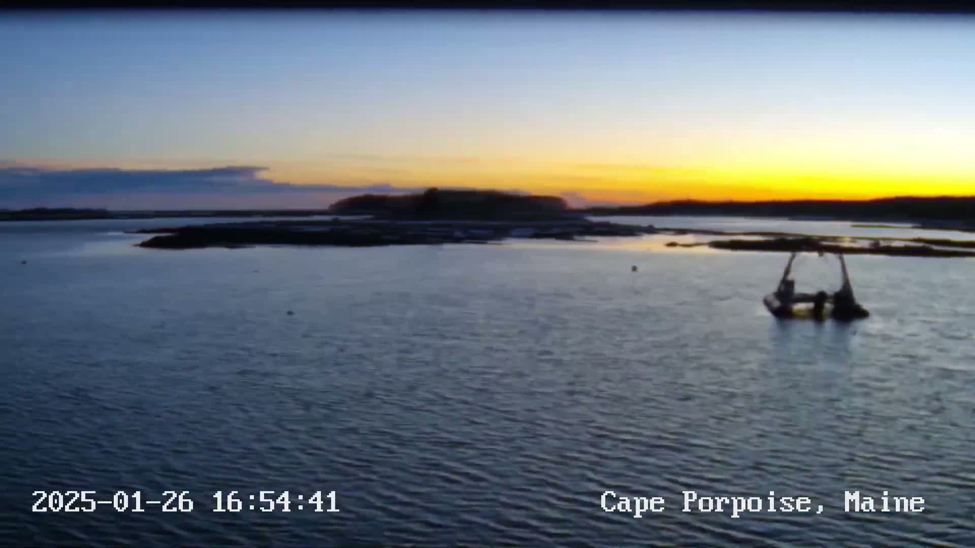 A calm body of water at sunset shows a small island and a vessel in the middle distance.
