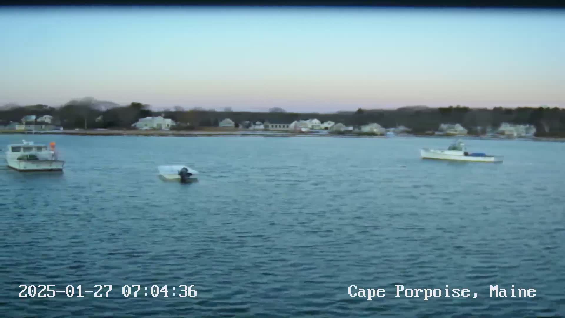 Three boats are moored in calm water in front of a shoreline with houses and trees under a pale, clear sky.
