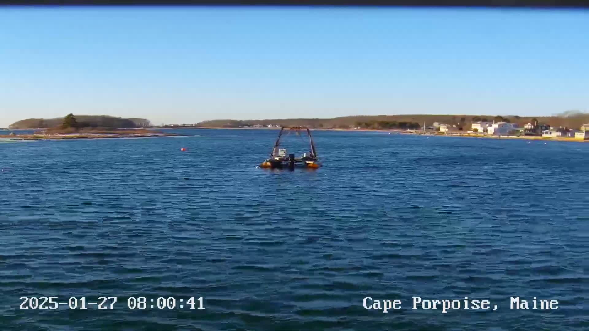 A small boat with a metal frame sits in calm, blue water under a clear blue sky, with land visible in the distance.