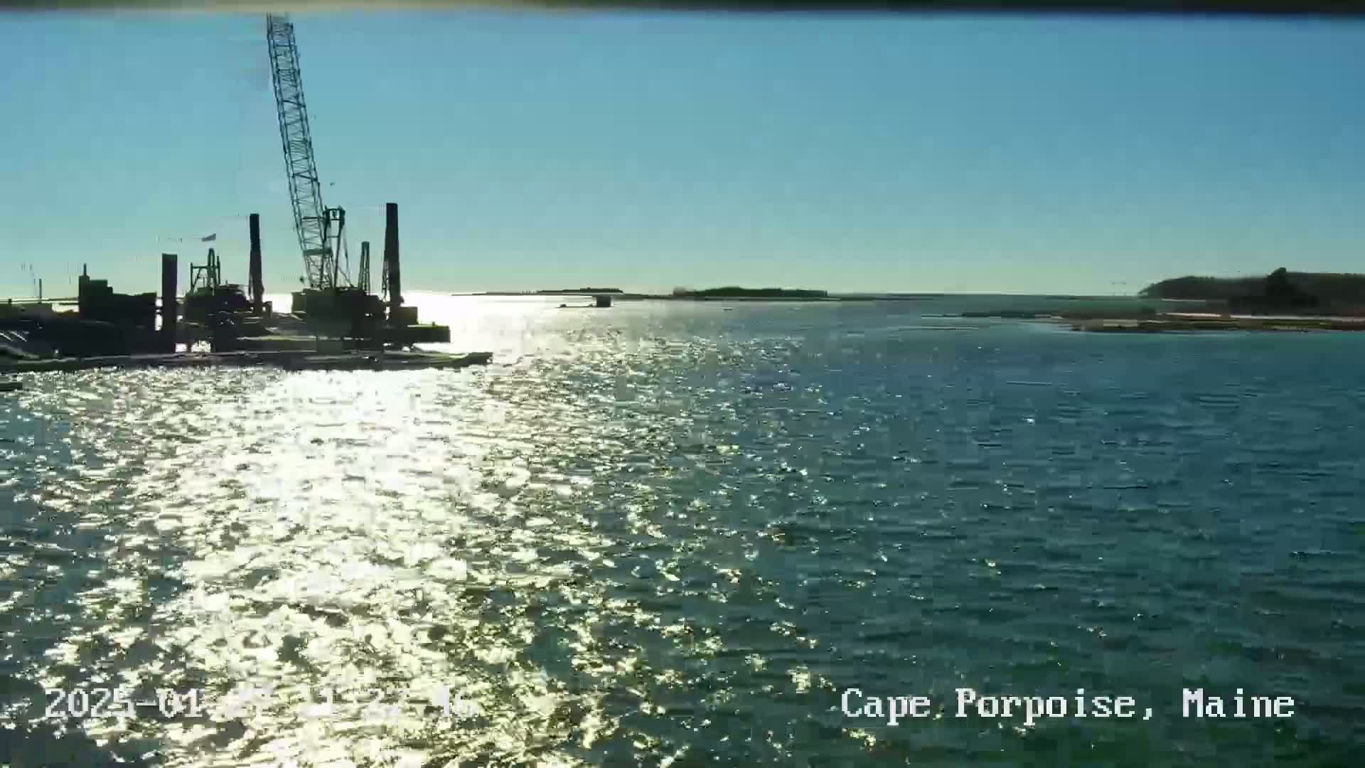 A large crane sits on a barge in calm, sunlit water near a shoreline under a clear blue sky.