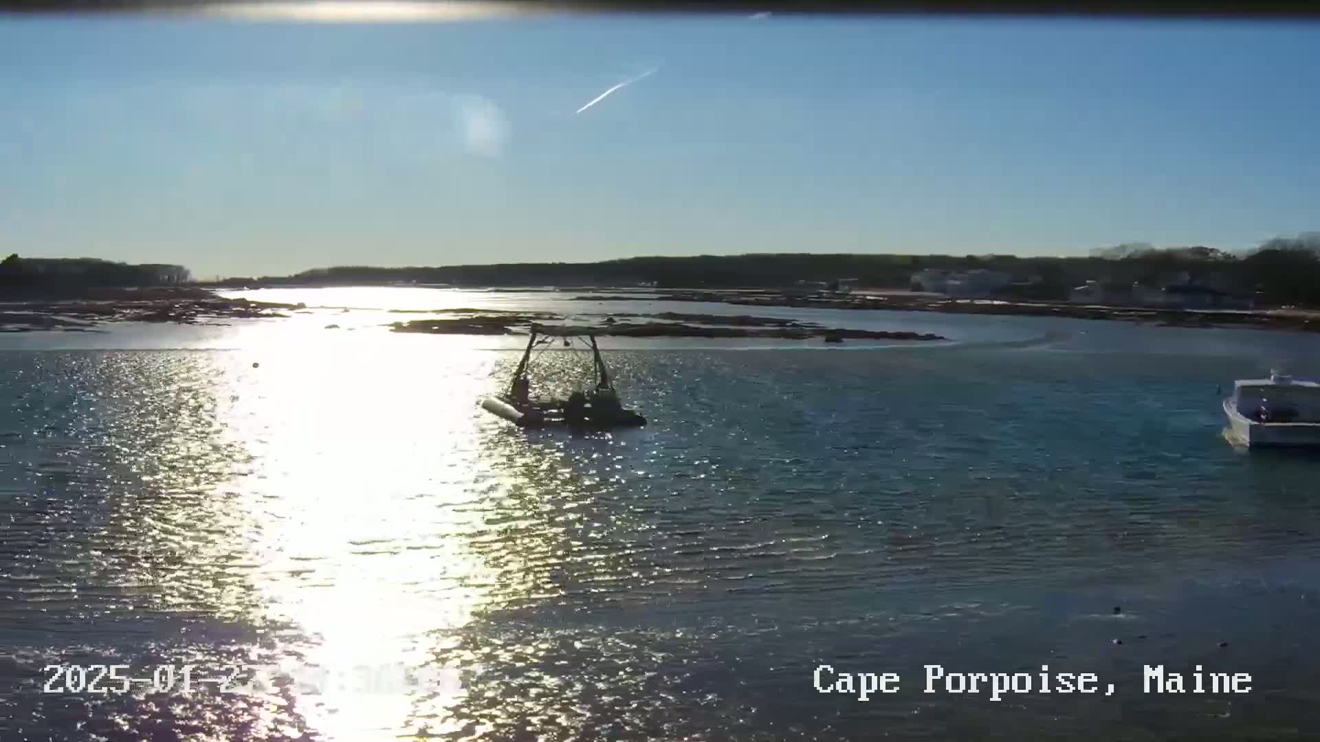 A sunny day shows a calm body of water reflecting sunlight, with a small boat and a larger vessel in the distance near a shoreline.