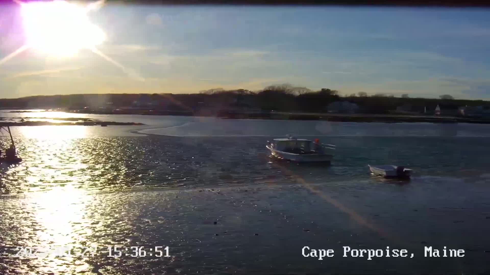 Two small boats are on calm water reflecting the bright sunset over a shoreline with trees and houses.