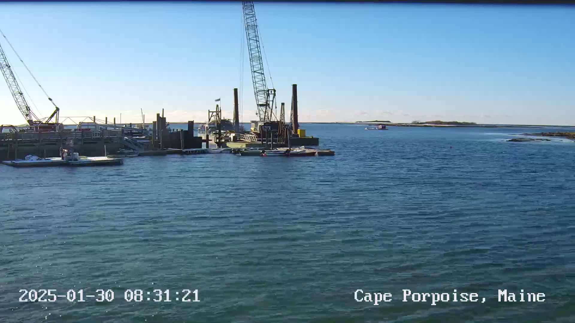 A construction barge with a crane is situated in a calm body of water under a clear blue sky.