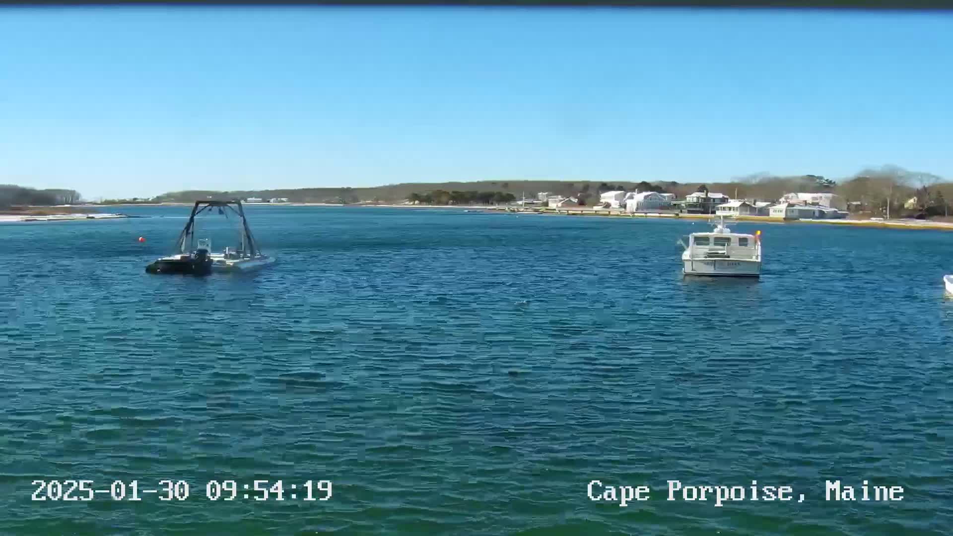 Under a clear blue sky, a body of water shows two small boats and a  larger vessel with a crane-like apparatus, near a shoreline with houses and trees.