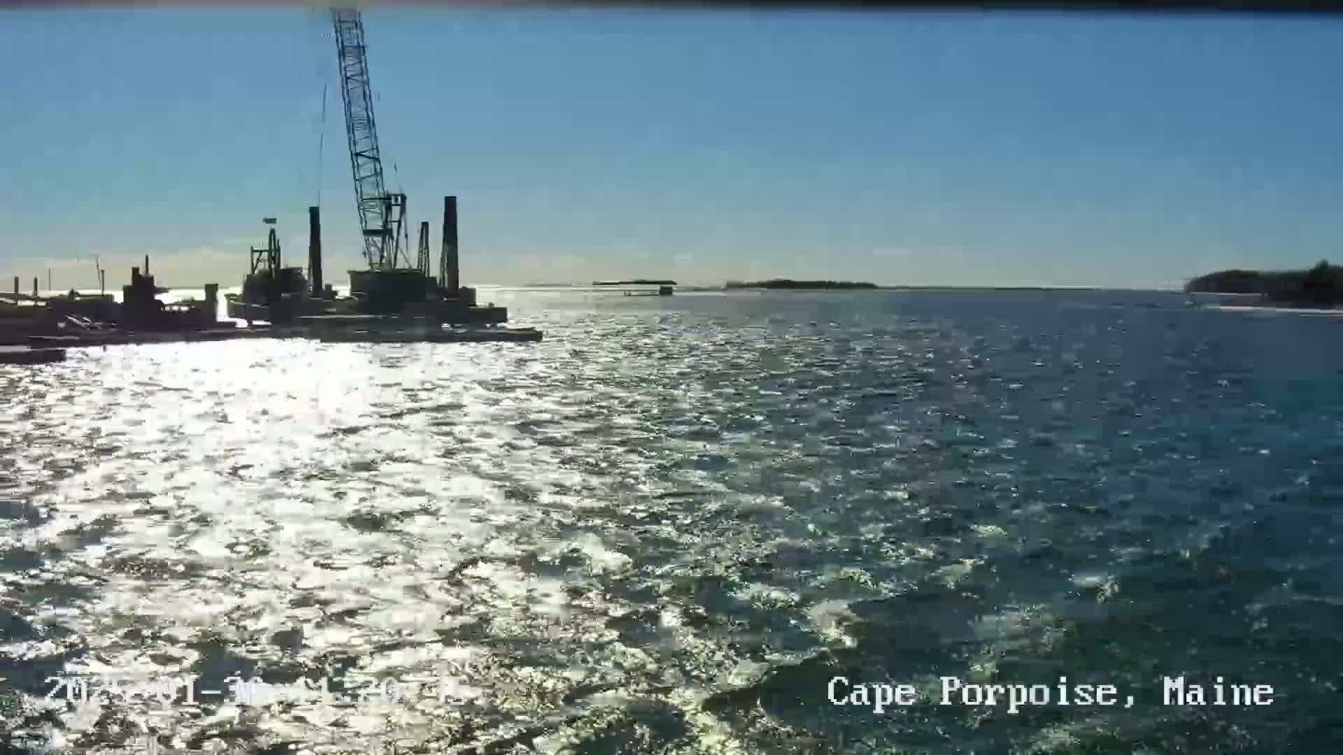 A large crane and other construction equipment are visible on a body of water under a sunny sky with a slight ripple on the surface.