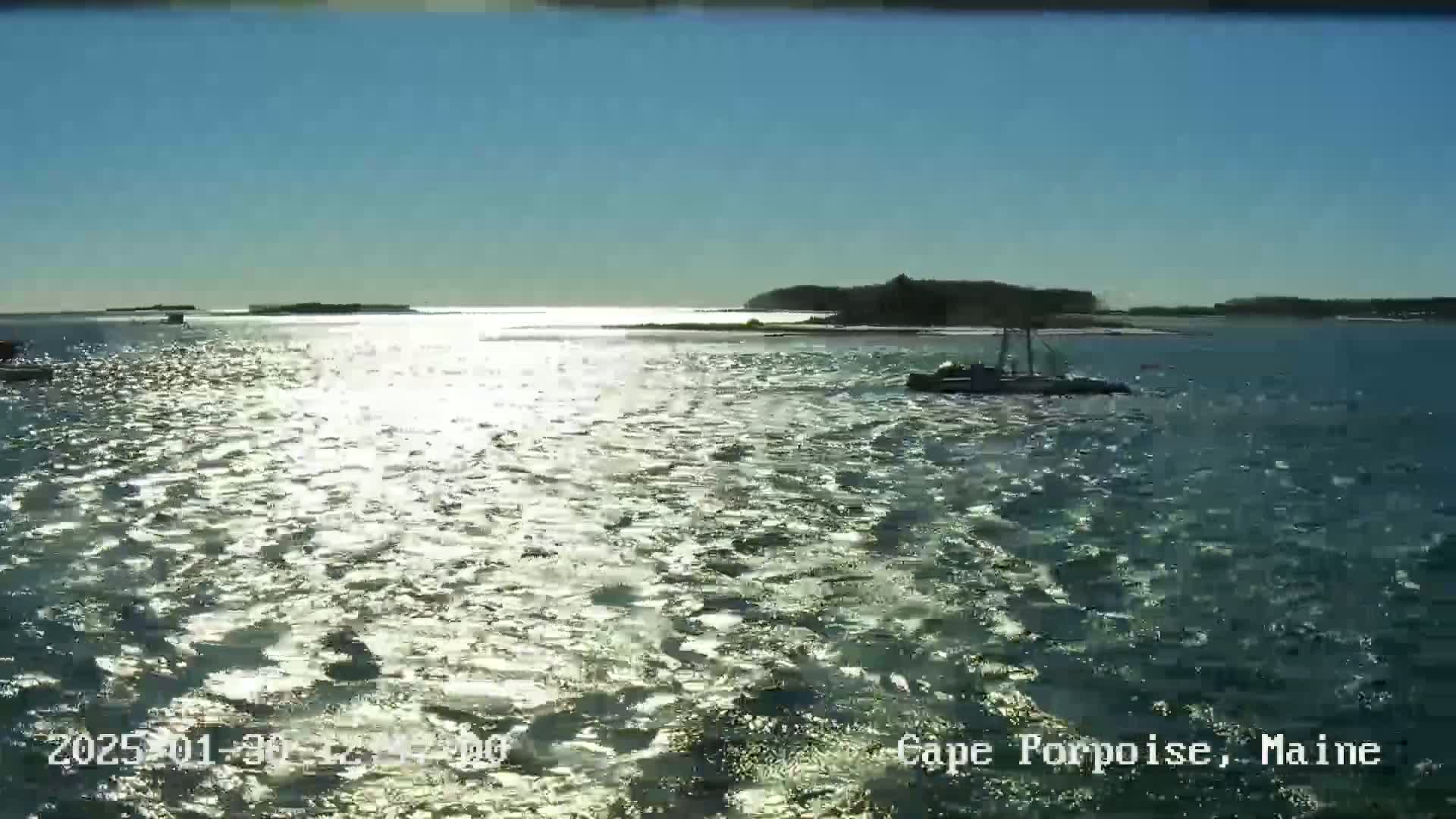 A sunny day shows a calm body of water reflecting sunlight, with a few boats and distant land visible.
