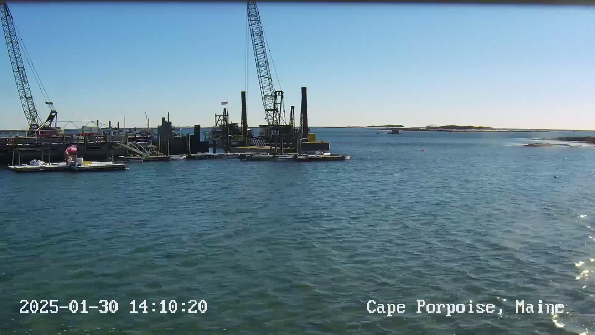 A body of water on a sunny day shows a dock with construction equipment, several small boats, and a distant shoreline.