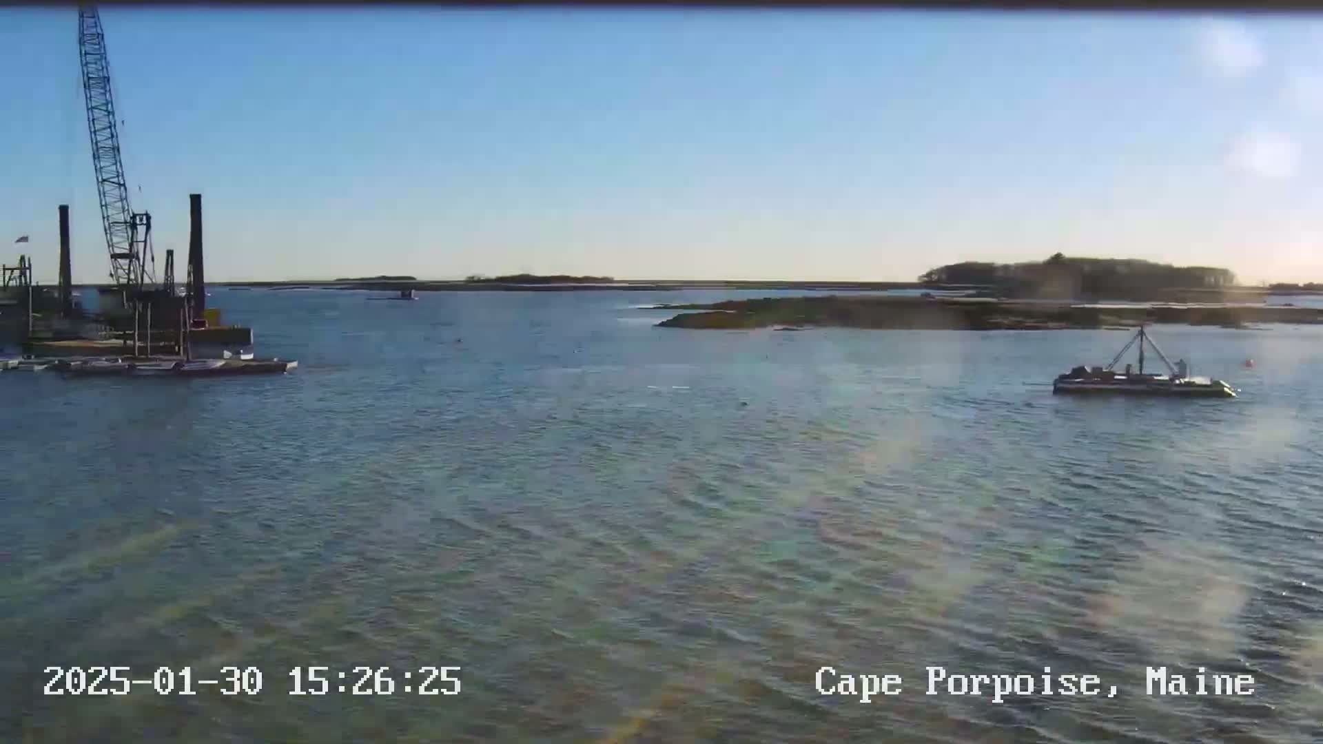 A calm body of water is shown with a crane and barge on the left, a small boat in the center, and land in the background under a clear, sunny sky.