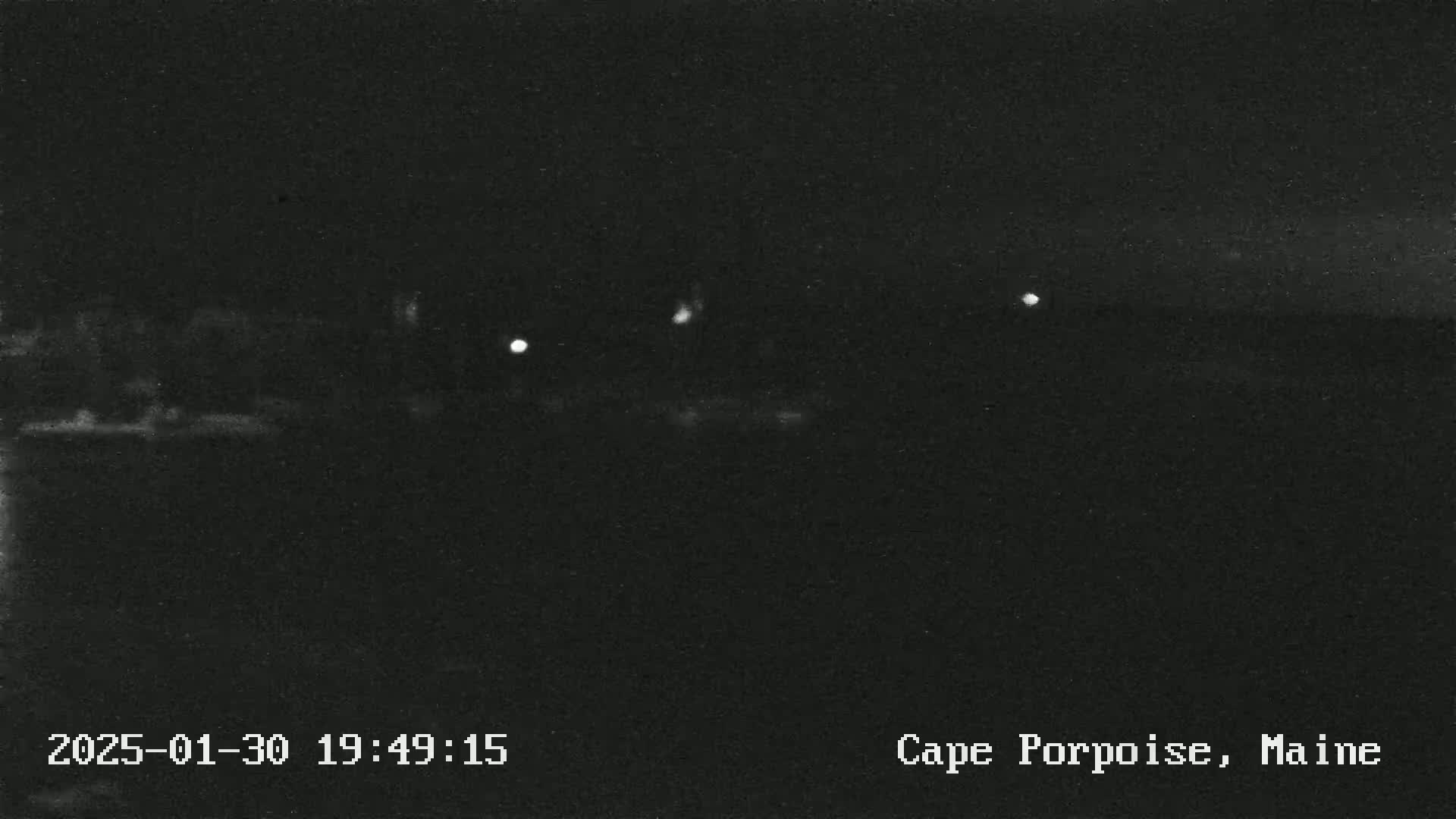 A nighttime, low-light image shows a dark body of water with a few distant lights visible on the horizon under a clear, dark sky.