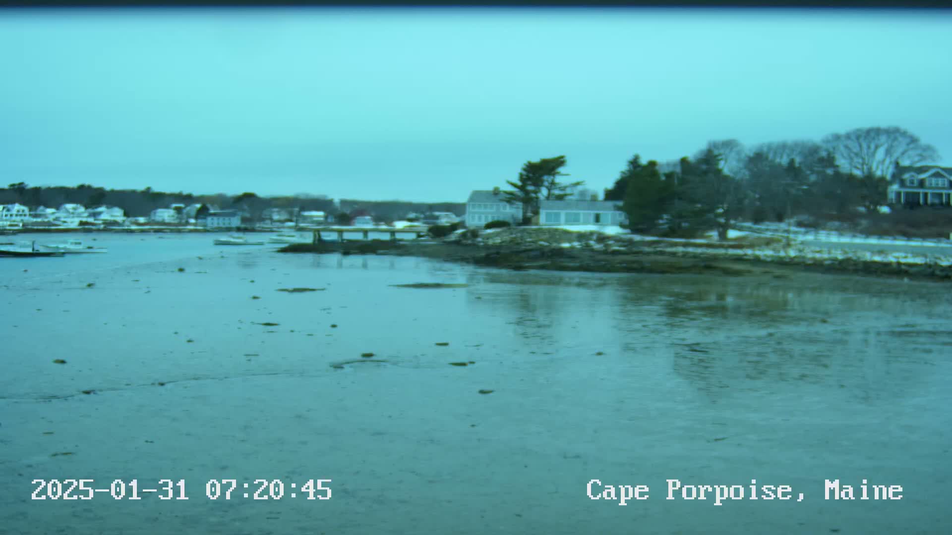 A calm, overcast day reveals a coastal scene with several houses and boats near the water's edge, some patches of snow are visible on the ground.