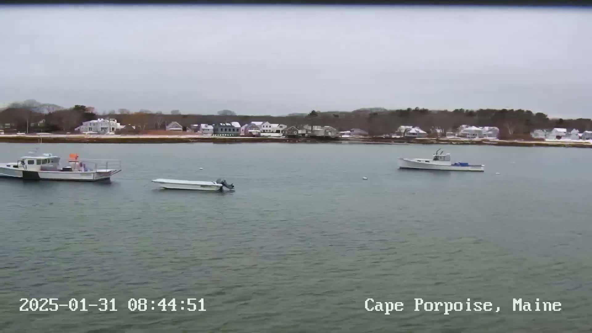 Three boats are moored in calm, gray water in front of a snow-dusted shoreline with houses and trees under an overcast sky.