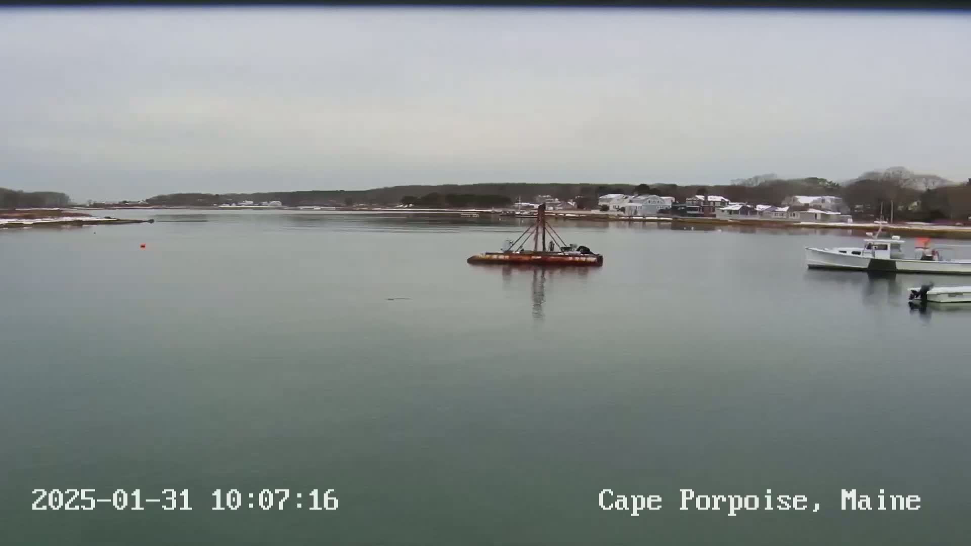 A calm, overcast day shows a body of water with a barge, two boats, and a snow-dusted shoreline in the distance.