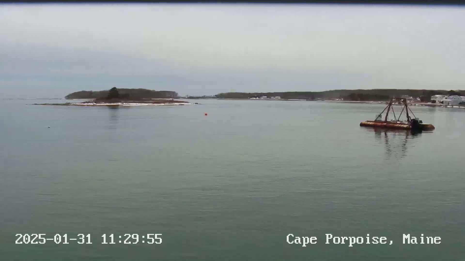 A calm, overcast day shows a small, snow-dusted island and a barge in a body of water.