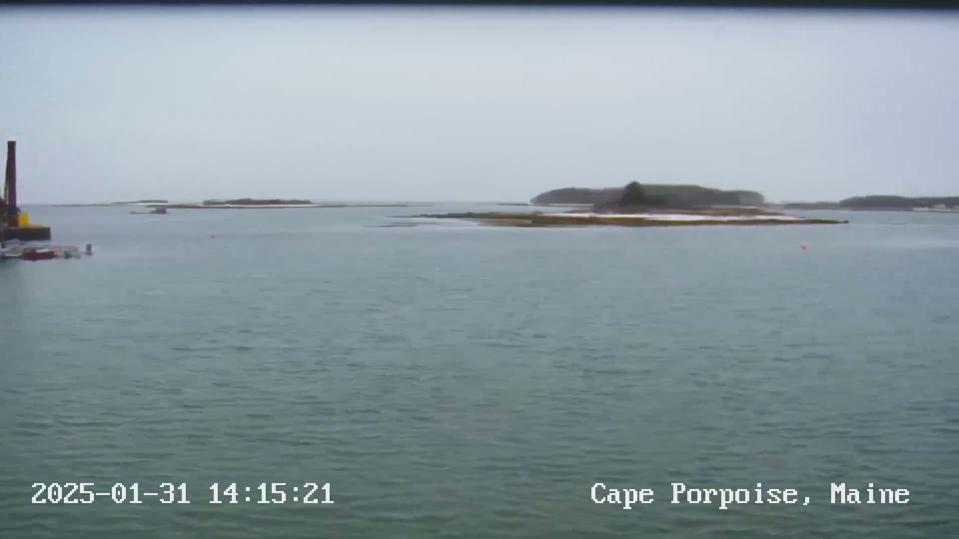 A calm body of water shows small islands with patches of snow under an overcast sky.