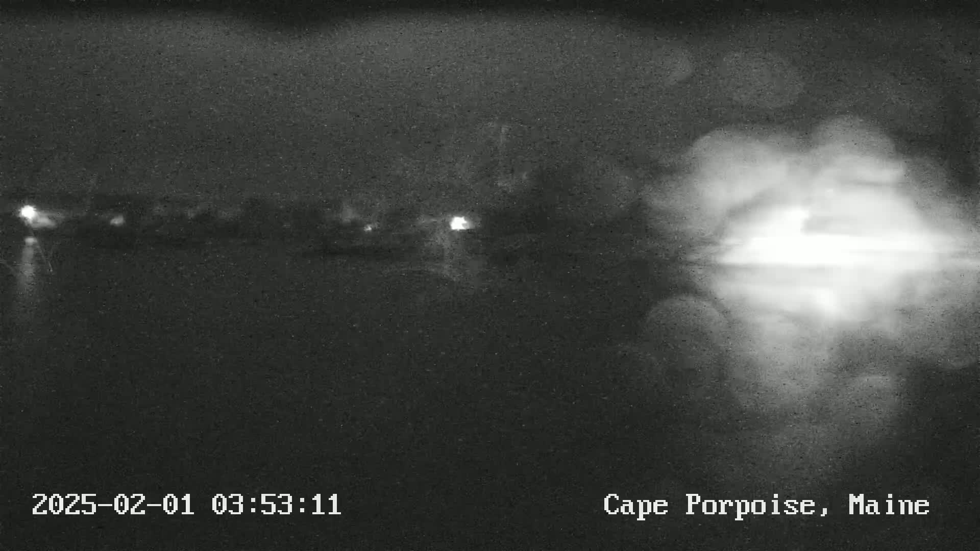 A nighttime, low-light image shows a blurry view of a body of water reflecting a bright light source, with faint lights visible in the distance across the water.
